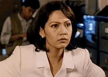 a woman in a white coat is sitting at a desk and looking at the camera with a serious look on her face .