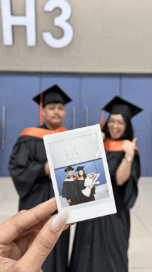 a person holding a picture of two graduates in front of a building with the number h3 on it