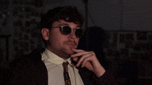 a man wearing sunglasses and a tie holds his finger to his chin