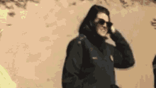 a woman wearing sunglasses and a black jacket is talking on a cell phone while standing in front of a wall .