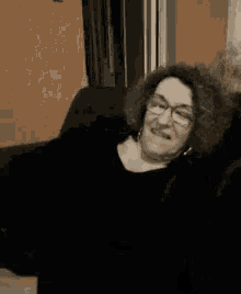 an elderly woman with curly hair and glasses is sitting on a couch and smiling .