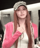 a woman with long red hair is wearing a pink jacket and a hat and giving a thumbs up