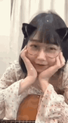 a girl wearing cat ears and glasses is holding her hands to her face while playing a guitar .