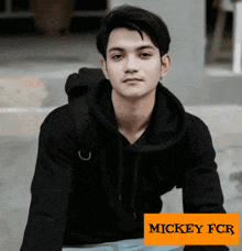 a young man wearing a black hoodie sits in front of a mickey fcr sign