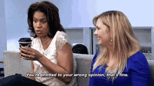 two women are sitting on a couch talking to each other and one of them is holding a cup of coffee .