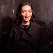 a woman wearing a black trench coat and a black headband is laughing
