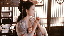 a woman in a kimono is sitting in a room with a window and a shelf .