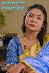 a woman in a blue and yellow saree is sitting at a table in front of a wall that says jo kahana he kaho