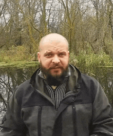 a man with a beard wearing a black jacket stands in front of a body of water