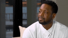 a man in a white shirt is sitting on a couch in front of a window and a screen that says ' interview '