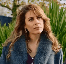 a woman wearing a fur coat and earrings is standing in front of a green bush .