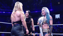 three women are standing in a wrestling ring and one of them is wearing a championship belt .