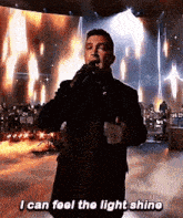 a man is singing into a microphone on a stage while saying `` i can feel the light shine '' .