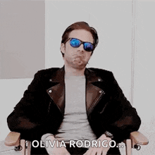 a man wearing sunglasses and a leather jacket is sitting in a chair and saying `` olivia rodrigo '' .