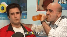 a man is drinking a bottle of orange juice while another man looks on .