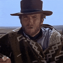a man with a beard wearing a hat and a poncho smoking a cigarette