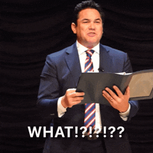 a man in a suit and tie is holding a book and says " what ? ! ? ! "
