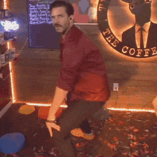 a man in a red shirt is squatting in front of a sign that says the coffee
