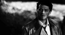 a black and white photo of a man in a trench coat and tie looking at the camera .