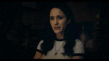a woman wearing a white t-shirt and a necklace looks at the camera in a dark room