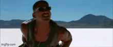 a man wearing sunglasses and a tank top is standing in the middle of a desert .