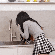 a woman in a plaid skirt is standing in front of a kitchen sink