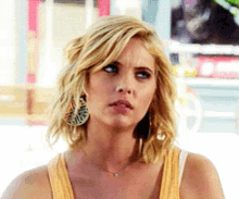 a blonde woman wearing a yellow tank top and earrings looks to the side