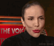 a woman with red lipstick is making a funny face in front of a red sign that says the voice .