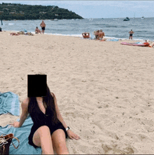 a woman sits on a beach with her face covered by a square