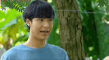 a young man in a blue shirt is making a funny face while standing next to a tree .