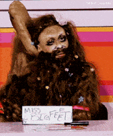a drag queen with a beard is sitting at a table with a sign that says miss bigfeet