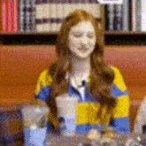 a woman in a yellow and blue striped sweater is sitting at a table in front of a bookshelf .