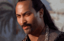 a man with long hair and a mustache is wearing a necklace .