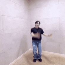 a man in a black shirt and blue jeans is dancing in a room with a white wall .