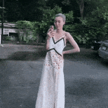 a woman in a long white dress is standing in a parking lot holding a cell phone .