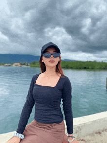 a woman wearing sunglasses and a hat sits in front of a body of water on a cloudy day