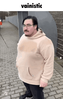 a man wearing glasses and a tan hoodie is standing on a brick sidewalk with the word vainistic above him