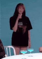 a woman in a black t-shirt is standing at a table with two glasses of blue liquid .