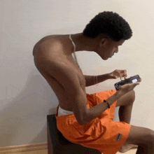 a shirtless man in orange shorts is sitting on a stool looking at his phone
