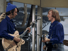 two men in blue shirts are playing guitars in front of a window