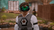 a man wearing a gray vest and green goggles is standing in front of a fence .