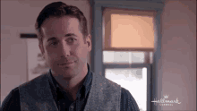 a man in a vest and shirt is standing in front of a window in a room .