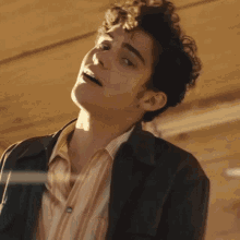 a young man with curly hair is wearing a brown jacket and a striped shirt