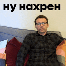 a man wearing glasses sits on a couch in front of a sign that says ну нахрен