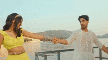 a man and a woman are holding hands in front of a body of water . the woman is wearing a yellow dress .