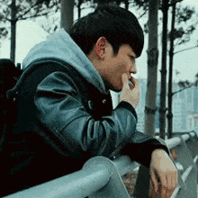 a young man in a leather jacket is leaning on a railing and smoking a cigarette .