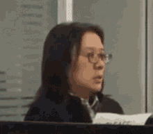 a woman wearing glasses sits at a desk with a piece of paper in front of her