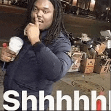 a man with dreadlocks is holding his finger to his lips while holding a cup of coffee .