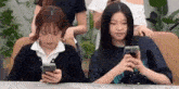 two women are sitting at a table looking at their cell phones .
