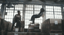 a man is standing on top of a pile of cardboard boxes while another man is doing a flip .